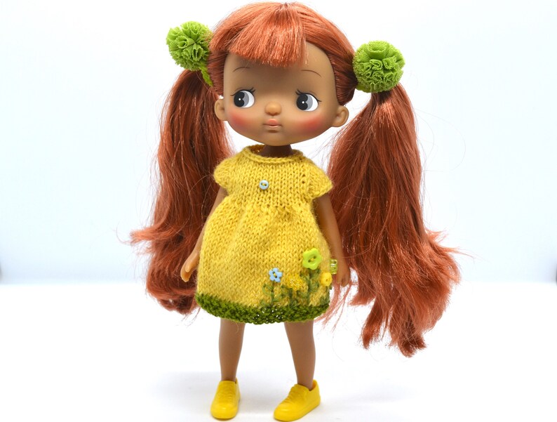 May include: A doll with long, red hair wearing a yellow knitted dress with green trim and floral accents. The doll has brown eyes and brown skin.