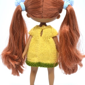 May include: A doll with long, auburn hair tied back with blue scrunchies. The doll is wearing a yellow knitted dress with a green trim and blue shoes.