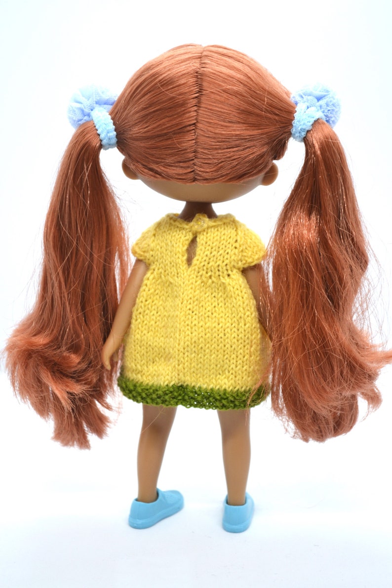 May include: A doll with long, auburn hair tied back with blue scrunchies. The doll is wearing a yellow knitted dress with a green trim and blue shoes.
