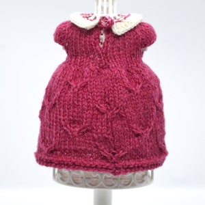 May include: A knitted magenta dress with a white collar and a small pink rose detail. The dress is displayed on a white mannequin. The dress has a textured pattern and short sleeves.