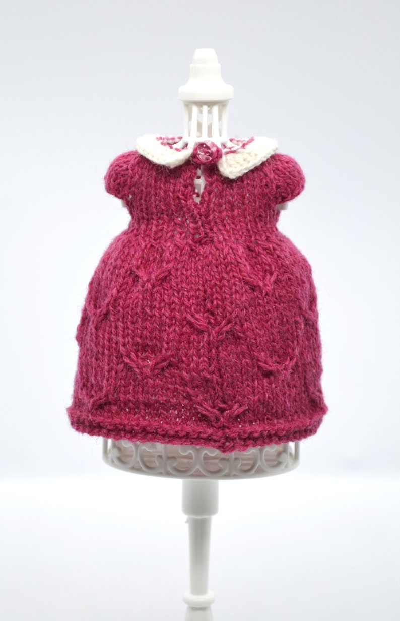 May include: A knitted magenta dress with a white collar and a small pink rose detail. The dress is displayed on a white mannequin. The dress has a textured pattern and short sleeves.