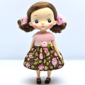 May include: A brown and pink floral dress with a pink knitted sweater top and pink flower accents. The doll has brown hair and a light complexion.
