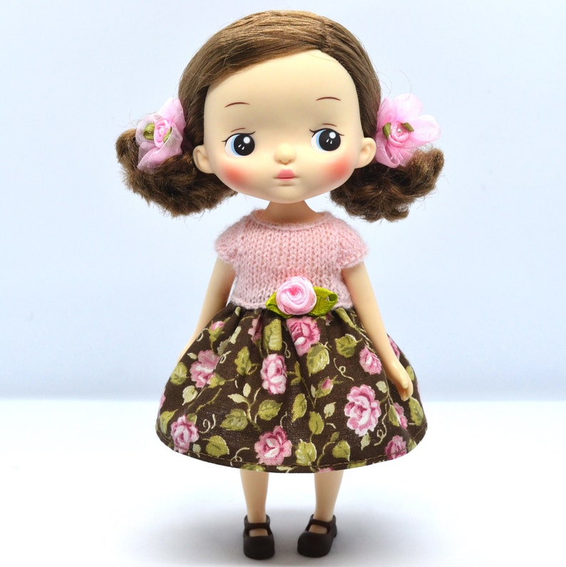 May include: A brown and pink floral dress with a pink knitted sweater top and pink flower accents. The doll has brown hair and a light complexion.