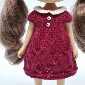May include: A knitted doll dress in a deep red colour, featuring a white collar and two white buttons. The dress has short sleeves and decorative pockets. The doll has long brown hair. The dress is designed for a doll.