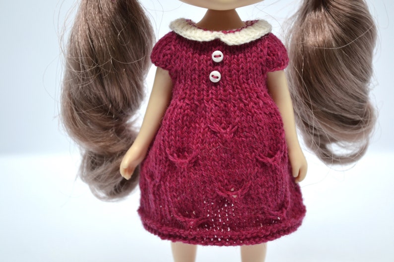 May include: A knitted doll dress in a deep red colour, featuring a white collar and two white buttons. The dress has short sleeves and decorative pockets. The doll has long brown hair. The dress is designed for a doll.