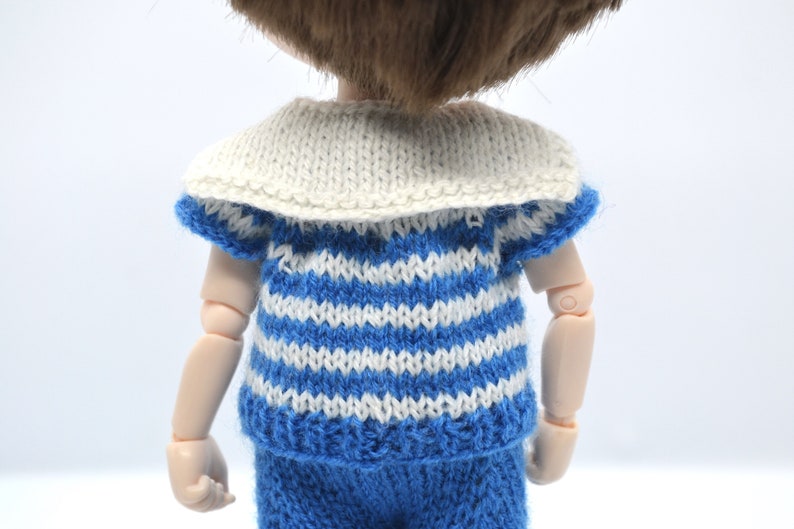 May include: A doll wearing a blue and white striped knitted shirt with a white sailor collar and matching blue pants.