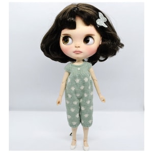 May include: A doll with dark brown bobbed hair, a light green butterfly hair clip, and large brown eyes. The doll wears a light green knitted romper featuring a white star pattern and a single small white button at the center of the neckline.
