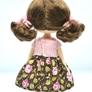 May include: A doll with brown hair in pigtails wearing a pink knitted cardigan and a brown floral skirt.