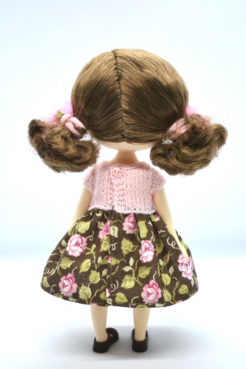 May include: A doll with brown hair in pigtails wearing a pink knitted cardigan and a brown floral skirt.