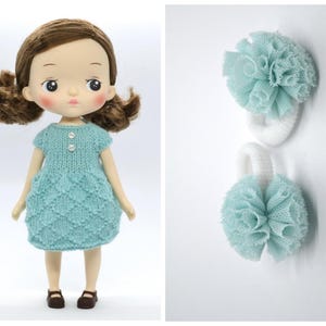 May include: A doll with brown hair wearing a light blue knitted dress and brown shoes. Two white hair ties with light blue pom-poms are also shown. The doll has rosy cheeks and is facing forward.