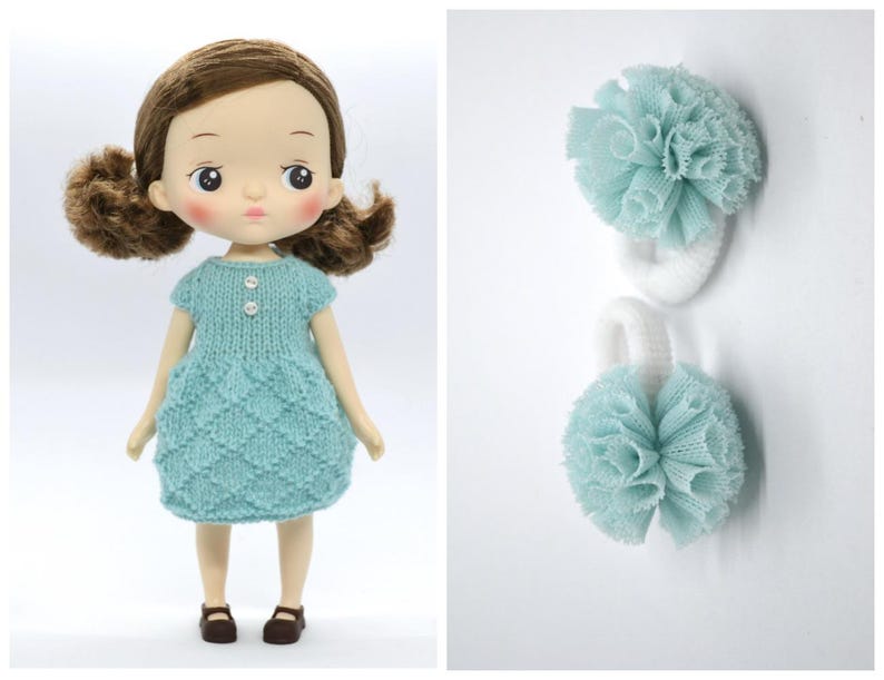 May include: A doll with brown hair wearing a light blue knitted dress and brown shoes. Two white hair ties with light blue pom-poms are also shown. The doll has rosy cheeks and is facing forward.