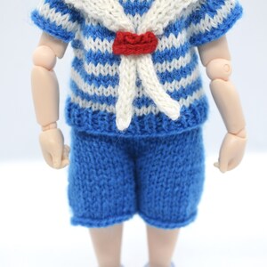 May include: A blue and white striped knitted sailor shirt with a white collar and a red bow tie. The shirt is paired with blue knitted shorts. The doll is wearing light purple shoes.