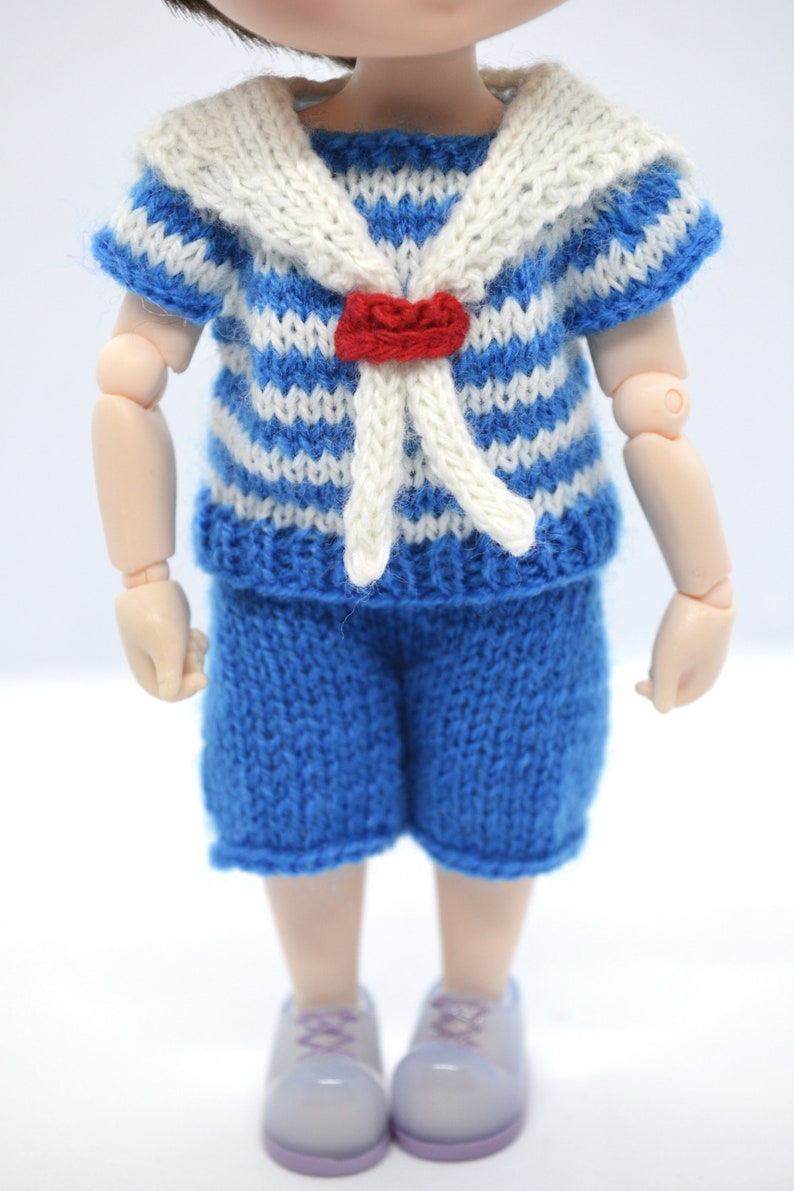 May include: A blue and white striped knitted sailor shirt with a white collar and a red bow tie. The shirt is paired with blue knitted shorts. The doll is wearing light purple shoes.