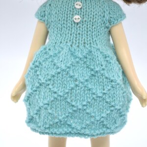 May include: A doll in a light blue knitted dress with a textured skirt and short sleeves. The dress features two white buttons at the neckline. The doll has brown hair and is set against a white backdrop.