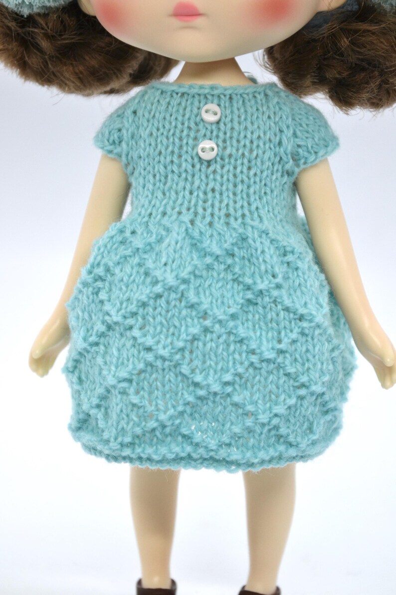 May include: A doll in a light blue knitted dress with a textured skirt and short sleeves. The dress features two white buttons at the neckline. The doll has brown hair and is set against a white backdrop.