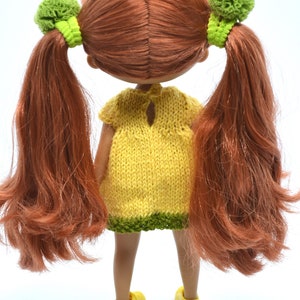 May include: A doll with long, red hair in pigtails with green bows. The doll is wearing a yellow knitted dress with a green trim and yellow shoes.