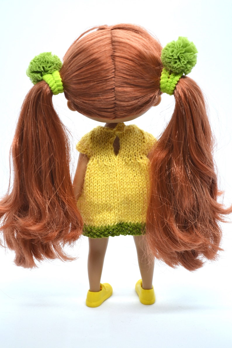 May include: A doll with long, red hair in pigtails with green bows. The doll is wearing a yellow knitted dress with a green trim and yellow shoes.