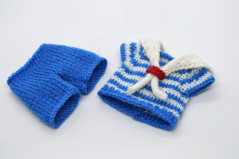 May include: A blue and white striped knitted sailor shirt with a white collar and a red button. The shirt is paired with blue knitted shorts.