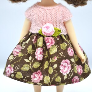 May include: A doll wearing a pink knitted top with a floral print skirt and a pink flower accent at the waist.