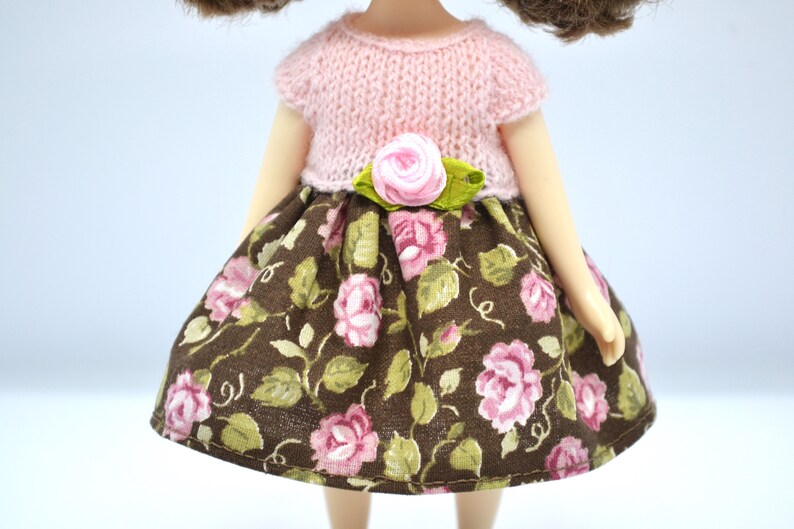 May include: A doll wearing a pink knitted top with a floral print skirt and a pink flower accent at the waist.