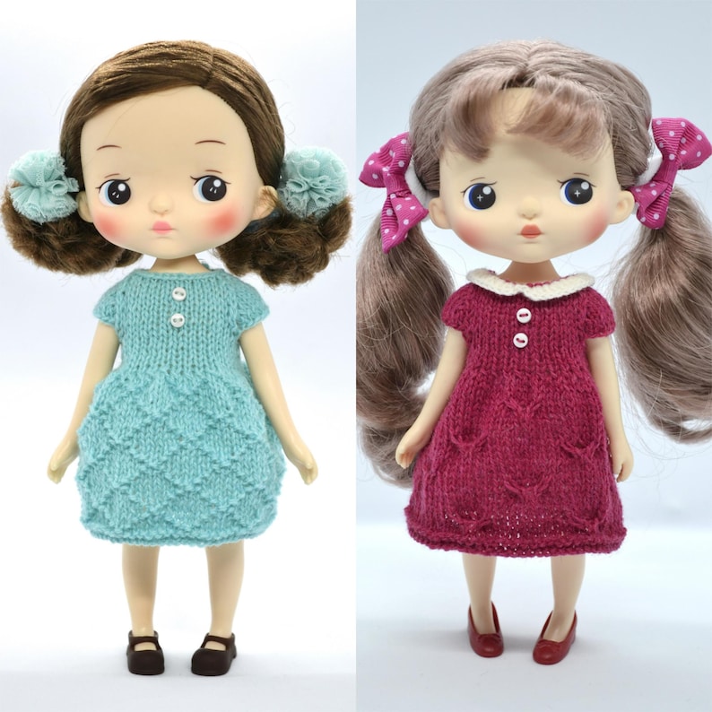 May include: Two dolls in knitted dresses. One doll has brown hair with blue pom-pom hair ties and a turquoise dress. The other doll has long brown pigtails with pink polka dot bows and a burgundy dress with a white collar.