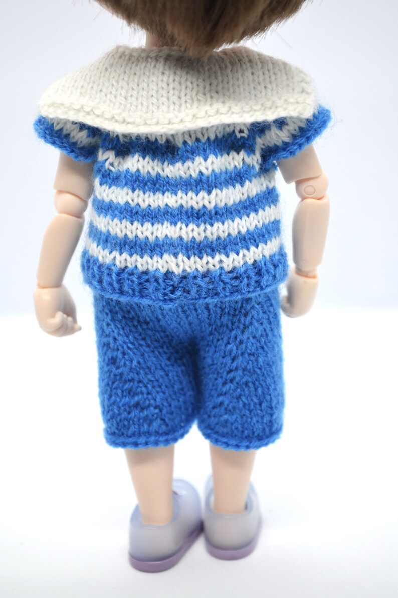 May include: A doll wearing a blue and white striped knitted shirt with a white sailor collar and blue knitted shorts.