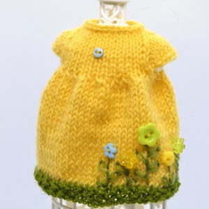 May include: A yellow knitted dress with green trim and embroidered flowers. The dress is on a white display stand.