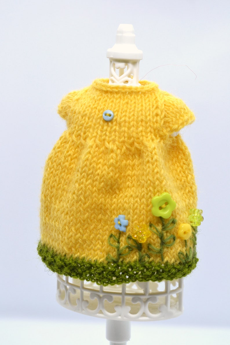 May include: A yellow knitted dress with green trim and embroidered flowers. The dress is on a white display stand.