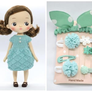 May include: A doll with brown hair wearing a light blue knitted dress and brown shoes. Beside the doll is a set of mint green hair accessories, including a headband with polka dots, hair clips, and barrettes. The words "Hand Made" are printed on the packaging.