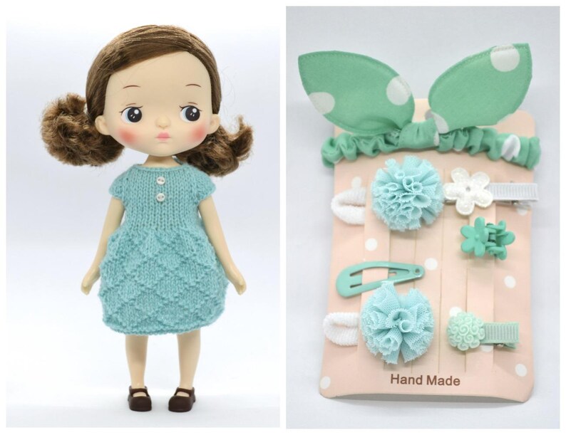 May include: A doll with brown hair wearing a light blue knitted dress and brown shoes. Beside the doll is a set of mint green hair accessories, including a headband with polka dots, hair clips, and barrettes. The words "Hand Made" are printed on the packaging.