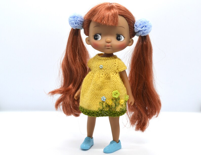 May include: A doll with brown hair and a yellow dress with green trim and blue flowers. The doll is wearing blue shoes.