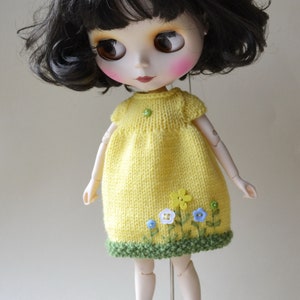 May include: A yellow knitted dress with green trim and embroidered flowers. The dress is worn by a doll with black hair and blue eyes. The doll is wearing white trainers.