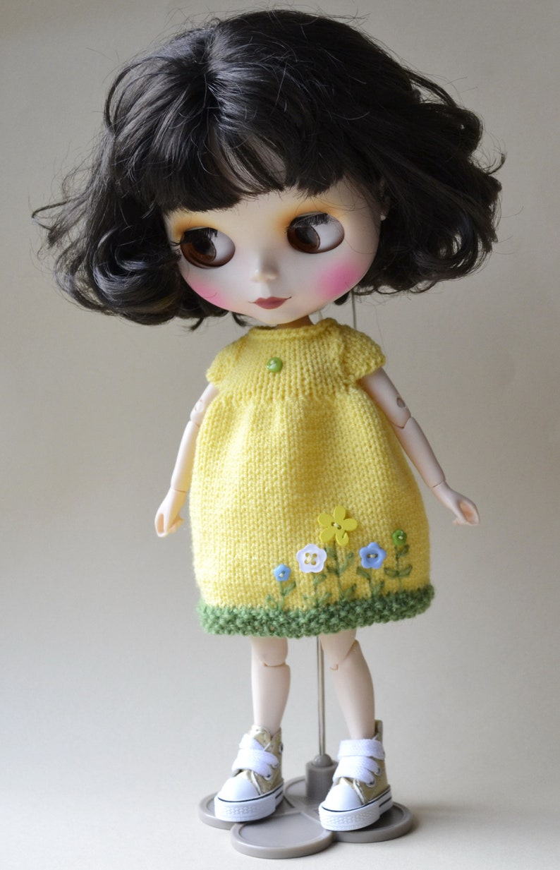 May include: A yellow knitted dress with green trim and embroidered flowers. The dress is worn by a doll with black hair and blue eyes. The doll is wearing white trainers.