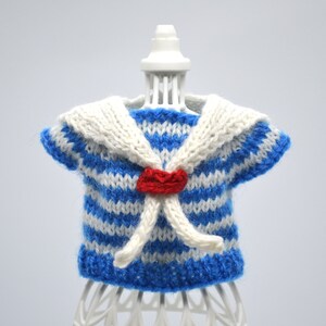 May include: A blue and white striped knitted sailor shirt with a white collar and a red bow. The shirt is on a white mannequin.