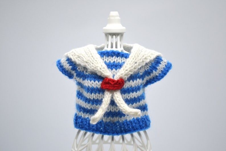 May include: A blue and white striped knitted sailor shirt with a white collar and a red bow. The shirt is on a white mannequin.