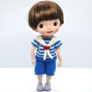 May include: A small doll with brown hair wearing a blue and white striped sailor shirt with a red bow and blue shorts.