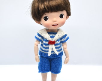 May include: A small doll with brown hair wearing a blue and white striped sailor shirt with a red bow and blue shorts.