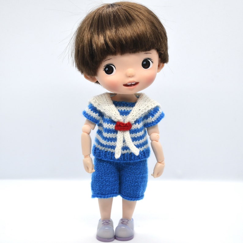 May include: A small doll with brown hair wearing a blue and white striped sailor shirt with a red bow and blue shorts.