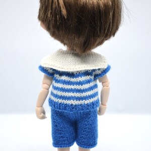 May include: A doll wearing a blue and white striped sailor shirt with a white collar and blue shorts. The doll has brown hair and is wearing light blue shoes.