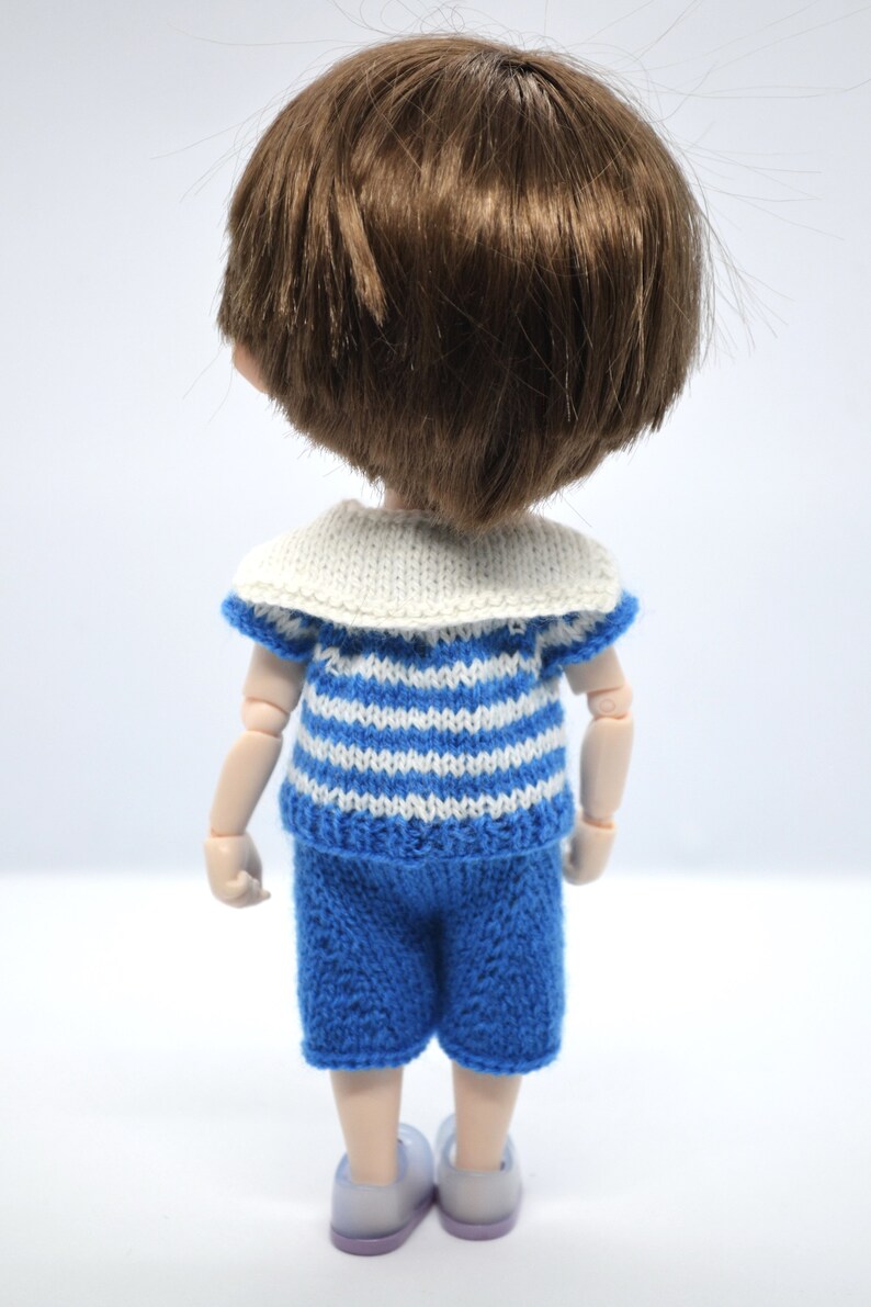 May include: A doll wearing a blue and white striped sailor shirt with a white collar and blue shorts. The doll has brown hair and is wearing light blue shoes.