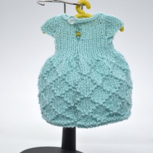 May include: A pale blue knitted dress with a textured skirt and short sleeves, displayed on a yellow hanger. The dress has two small buttons on the back and is presented on a black stand.