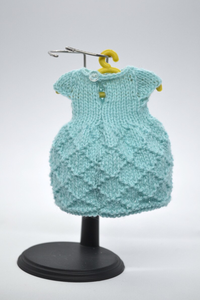 May include: A pale blue knitted dress with a textured skirt and short sleeves, displayed on a yellow hanger. The dress has two small buttons on the back and is presented on a black stand.