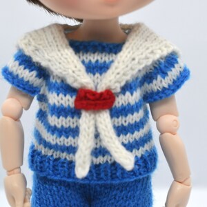 May include: A blue and white striped knitted shirt with a white sailor collar and a red bow tie. The shirt is designed for a doll.