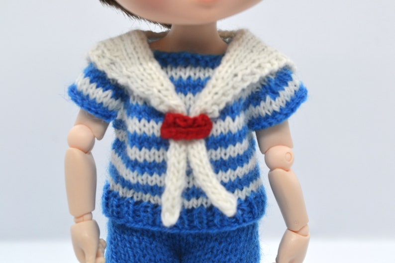 May include: A blue and white striped knitted shirt with a white sailor collar and a red bow tie. The shirt is designed for a doll.