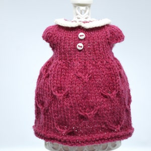 May include: A knitted magenta dress with a white collar and two white buttons. The dress is displayed on a white stand against a white background. The dress has a textured pattern.