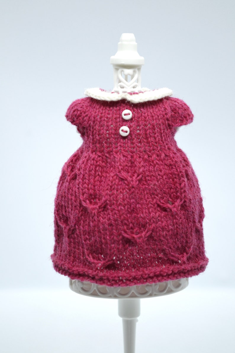 May include: A knitted magenta dress with a white collar and two white buttons. The dress is displayed on a white stand against a white background. The dress has a textured pattern.
