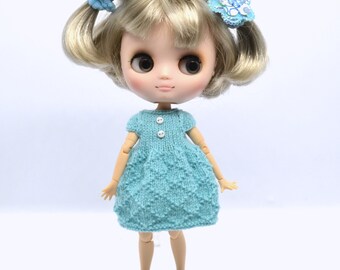 May include: A doll with blonde hair wearing a teal knitted dress with white buttons and blue shoes. The doll has blue flowers in her hair.