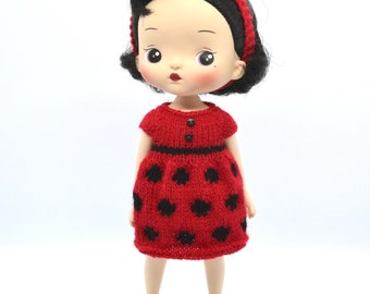 May include: A doll with black hair and a red headband with a ladybug detail. The doll wears a red knitted dress with black polka dots and a black belt. The doll has red shoes and a fair complexion.