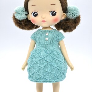 May include: A doll with brown hair in pigtails with blue accents. The doll is wearing a light blue knitted dress with two white buttons and brown shoes. The doll has rosy cheeks and a neutral expression.