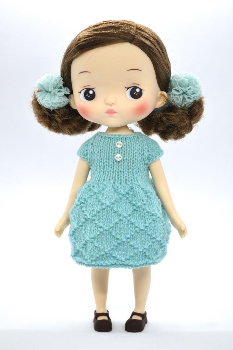 May include: A doll with brown hair in pigtails with blue accents. The doll is wearing a light blue knitted dress with two white buttons and brown shoes. The doll has rosy cheeks and a neutral expression.